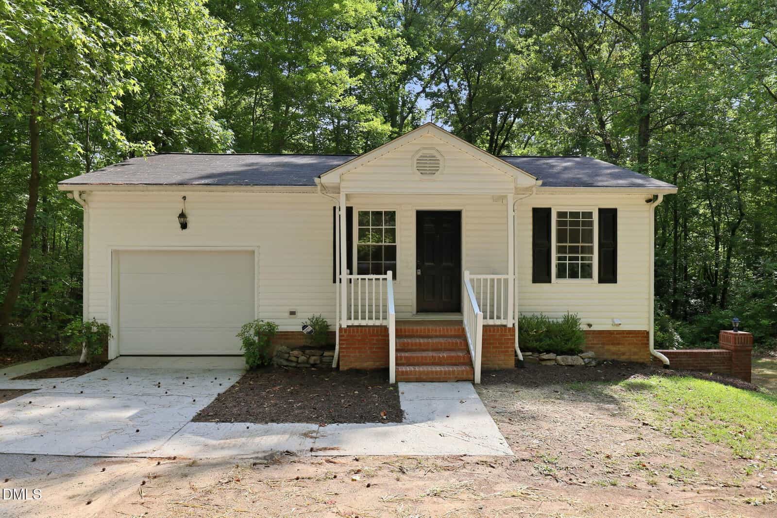 A small white house with black shutters and a black front door at 110 Fox Ridge Road, Louisburg, features front steps with white railings, a single-car garage, and is surrounded by green trees and shrubs.