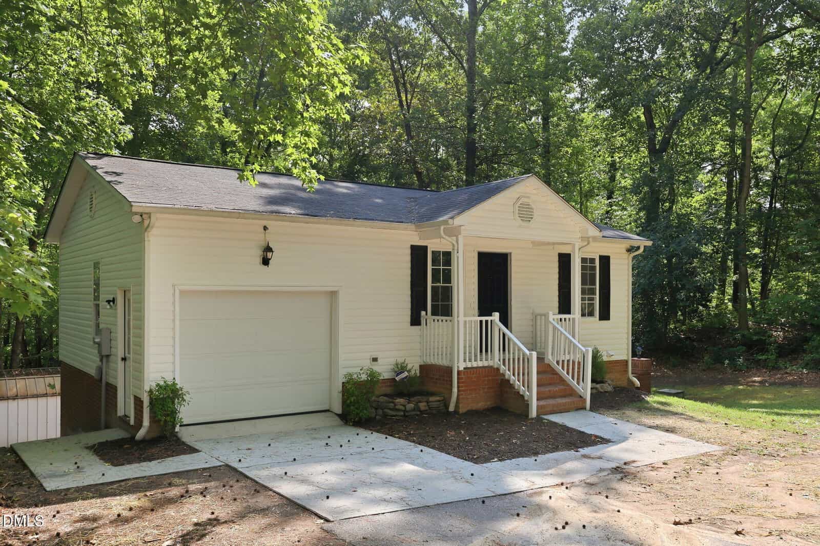 A small, single-story house with white siding, a one-car garage, black shutters, and a covered front porch with stairs, surrounded by trees and greenery on a sunny day.