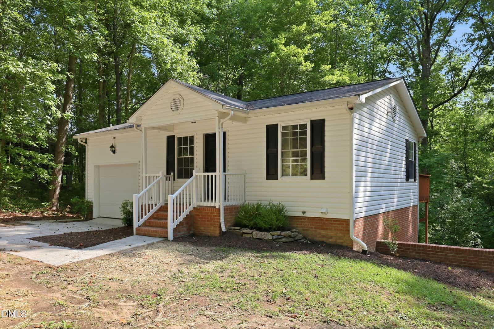 A small white house with black shutters, a covered front porch, and a single-car garage sits surrounded by trees. The yard features a sloped lawn and a concrete driveway.