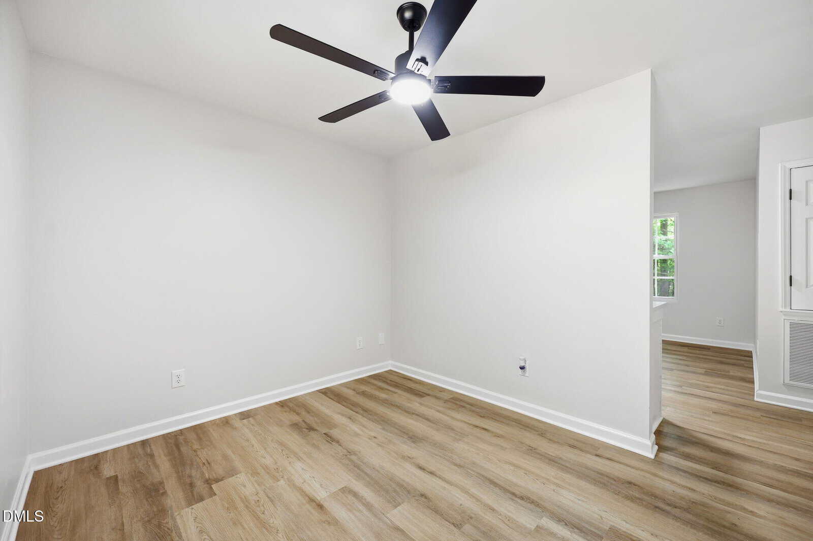A bright, empty room with light wood flooring, white walls, and a modern black ceiling fan with a light. An open doorway leads to another area with a window and more wood flooring.