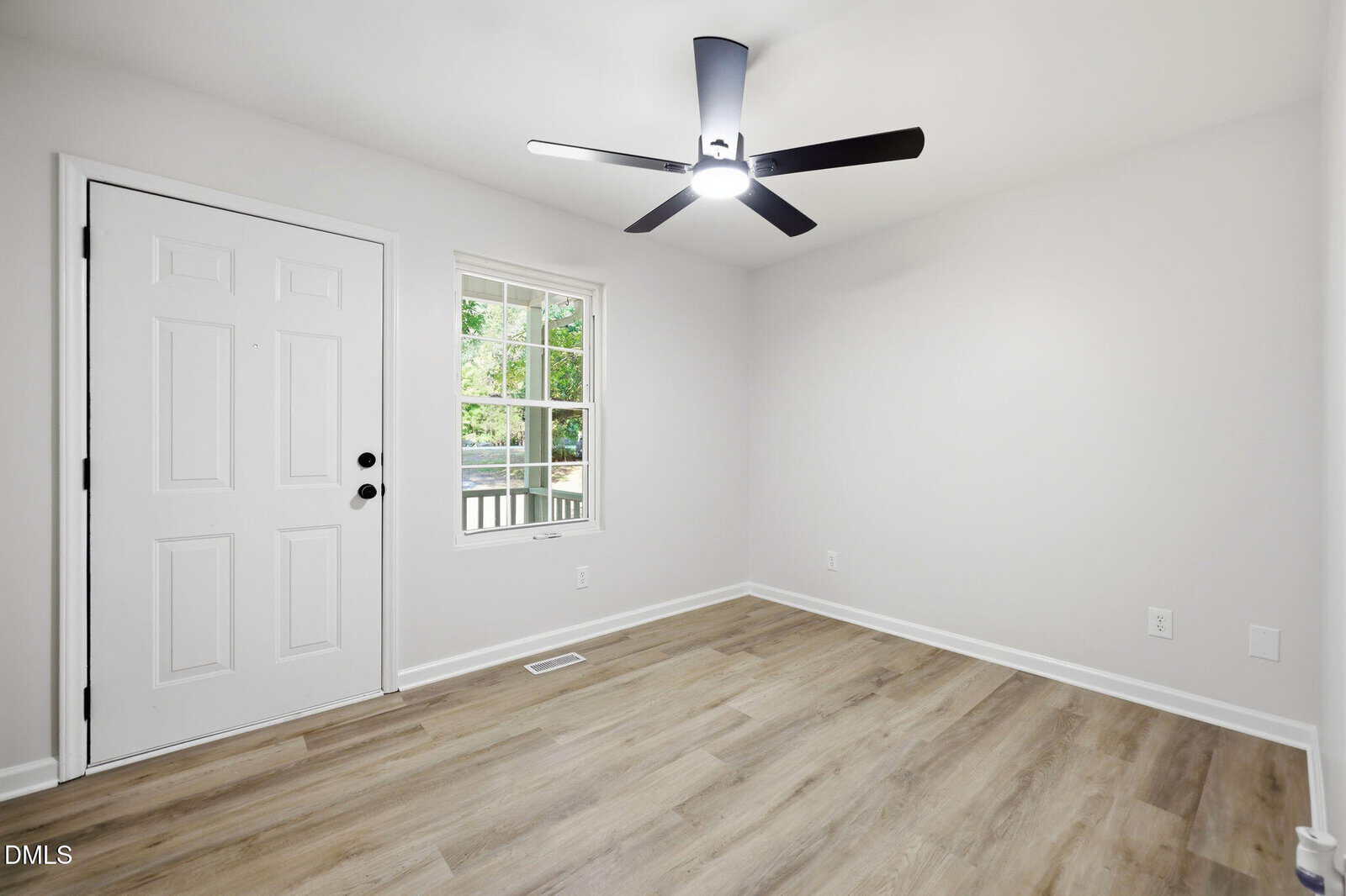 Bright, empty room with light wood flooring, white walls, a white door, a window with a view of greenery, and a modern black ceiling fan with a light.