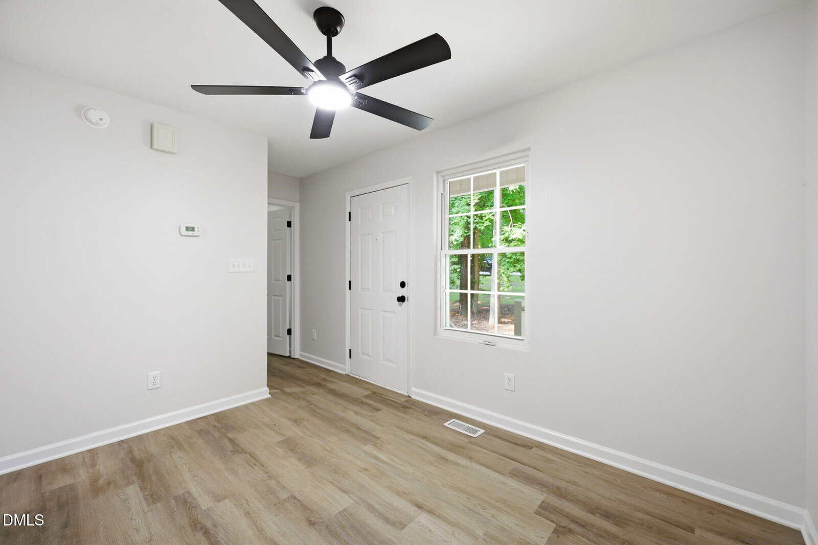 A bright, empty room with light wood flooring, white walls, a ceiling fan with black blades, a window showing greenery, and two white doors.