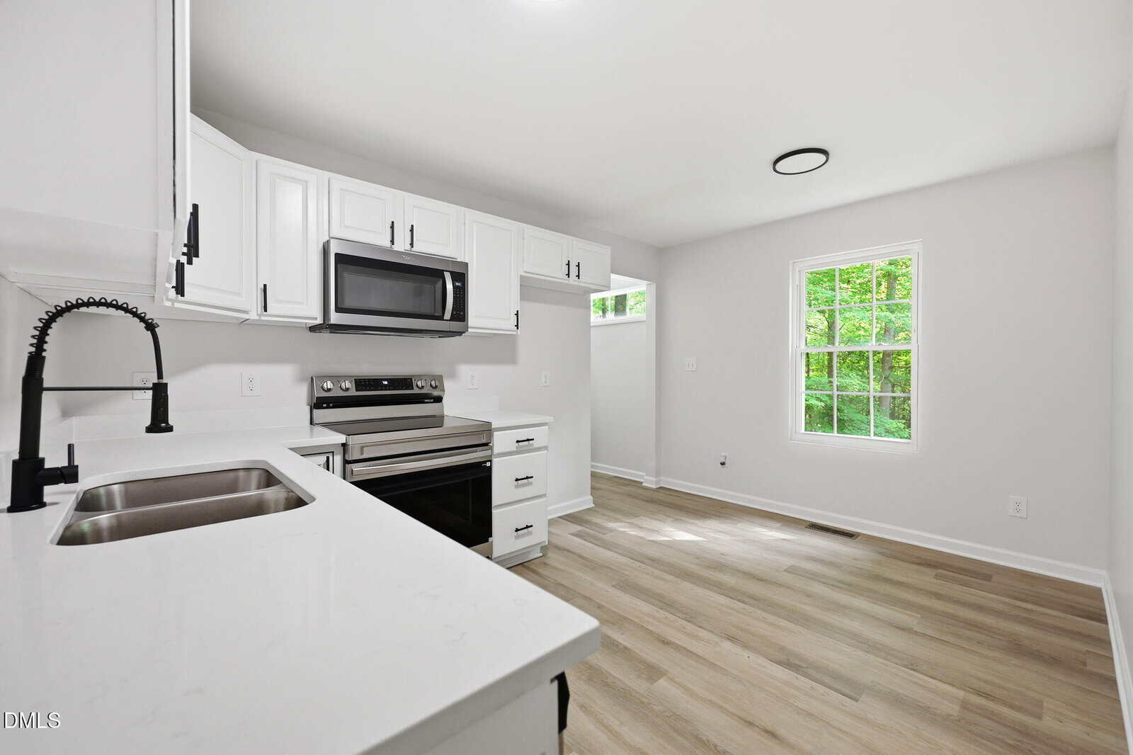 Modern kitchen with white cabinets, stainless steel appliances, light wood floors, a large window with green trees outside, and a black pull-down faucet. The room is bright and minimally decorated.