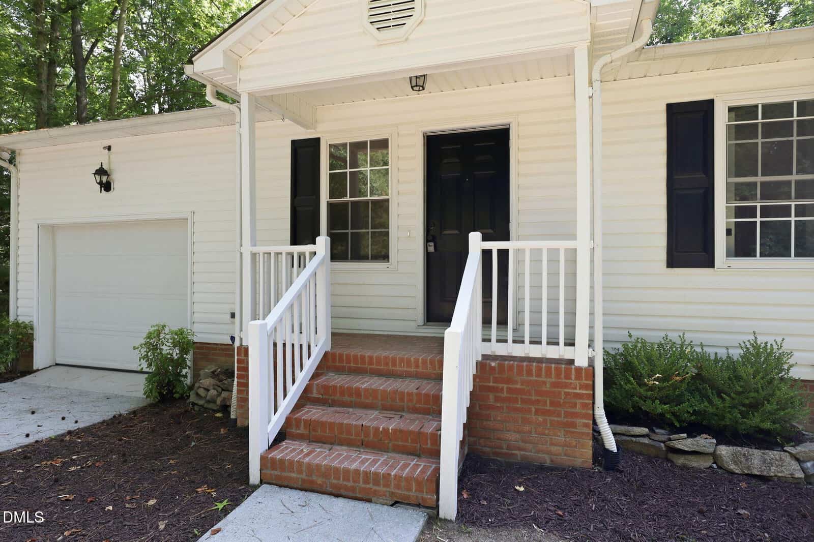 Front view of a white house with brick steps leading to a small porch with white railings, a black front door, and windows on either side. There are shrubs and mulch along the walkway.