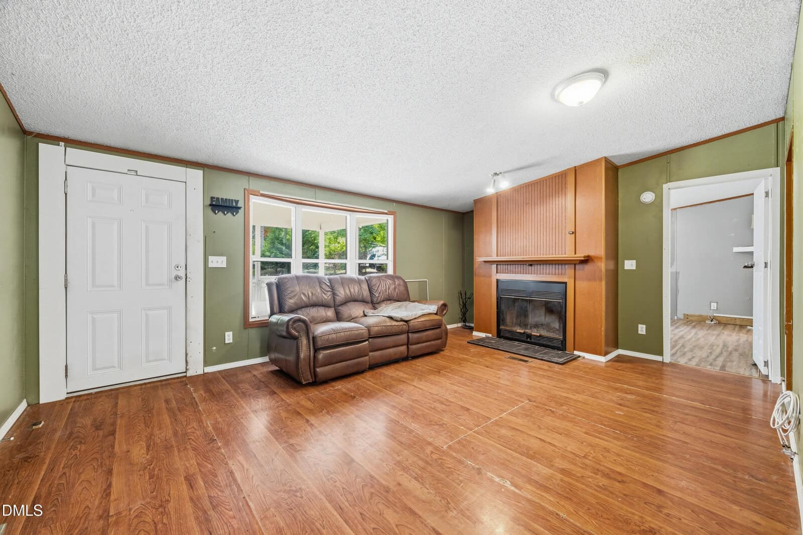 Living room with wood floor, green walls, a brown sofa by a window, a fireplace with a wood mantle, and a white front door. An adjacent room with lighter flooring is partially visible.