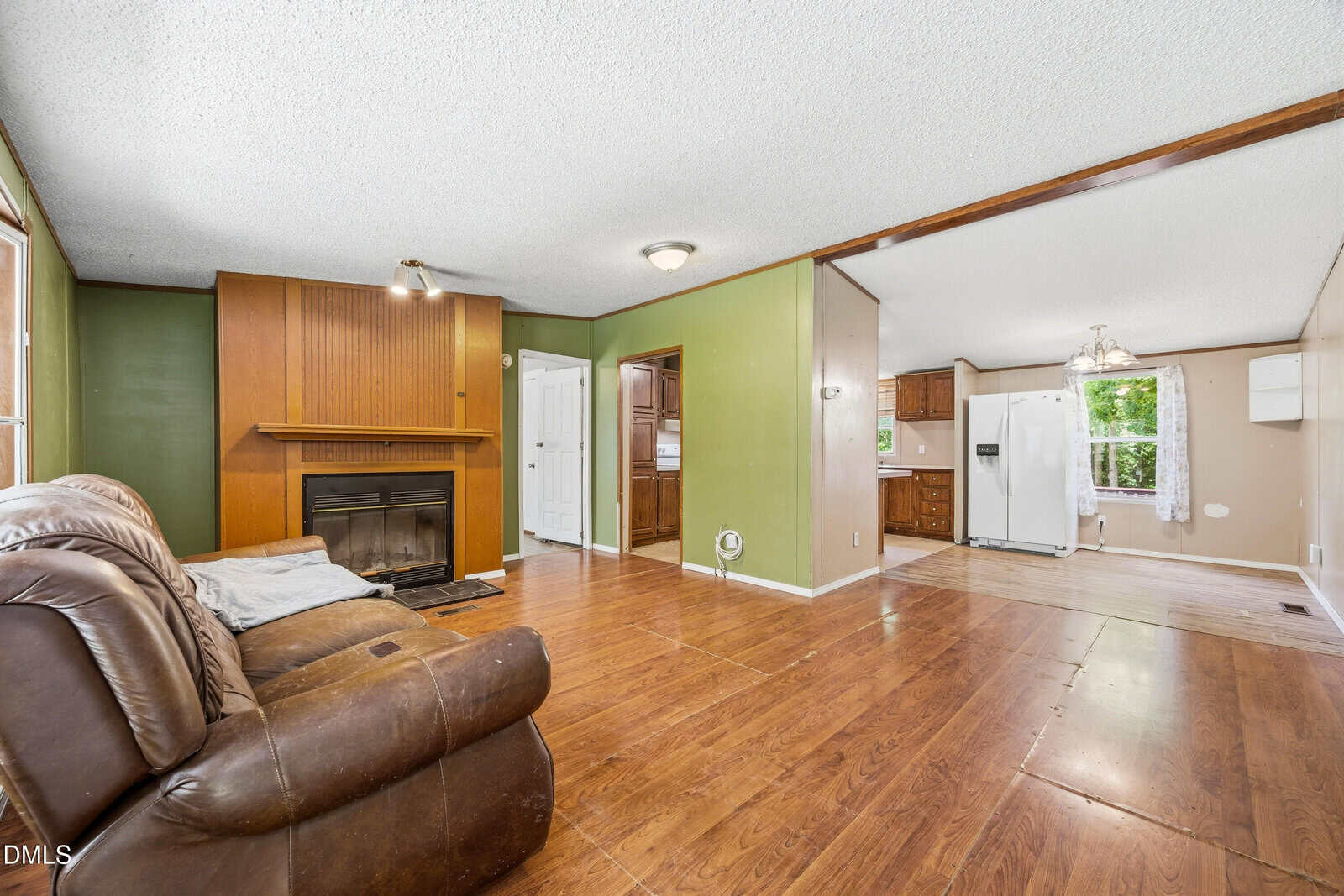 Living room with brown recliners, wood floors, green walls, fireplace, ceiling fan, and open view into a kitchen with white refrigerator and wooden cabinets. Large windows provide natural light.