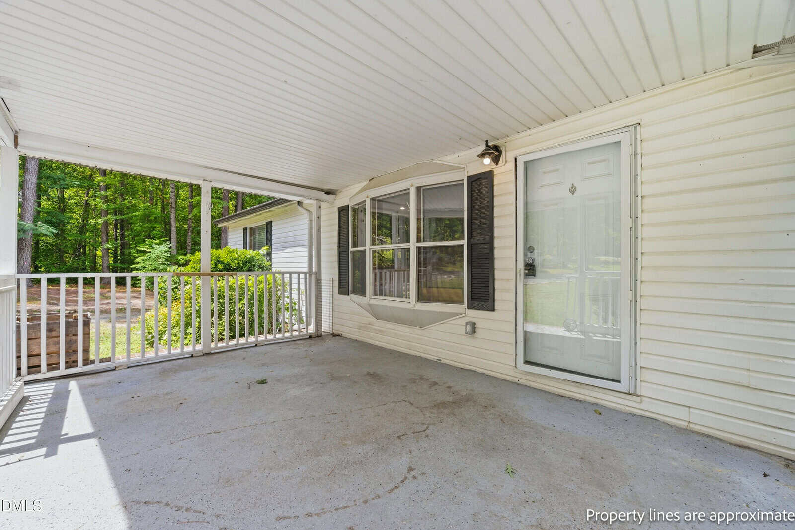 Covered front porch with concrete floor, white railing, beige siding, and a glass door with a side light. Large window with shutters faces a wooded yard. The space is clean and shaded.