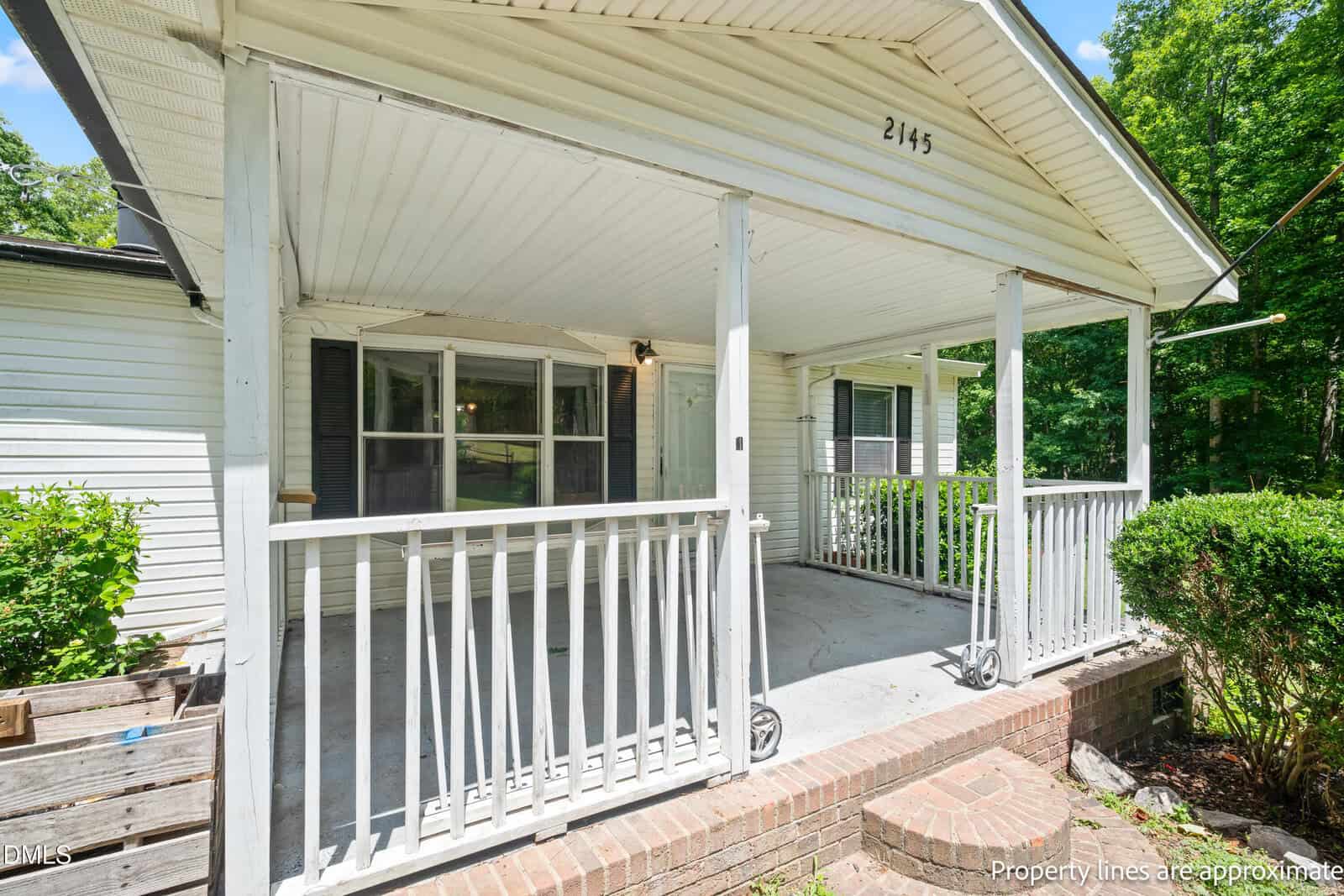 A white house with a covered front porch, white railings, and brick steps. Large windows face the porch, and green bushes and trees surround the property on a sunny day. The house number 2145 is visible.