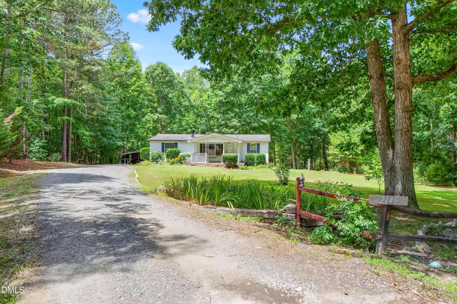 A white house with a front porch sits among tall trees and green lawn, with a gravel driveway leading to it and a red wooden fence beside the entrance displaying the number 2145.