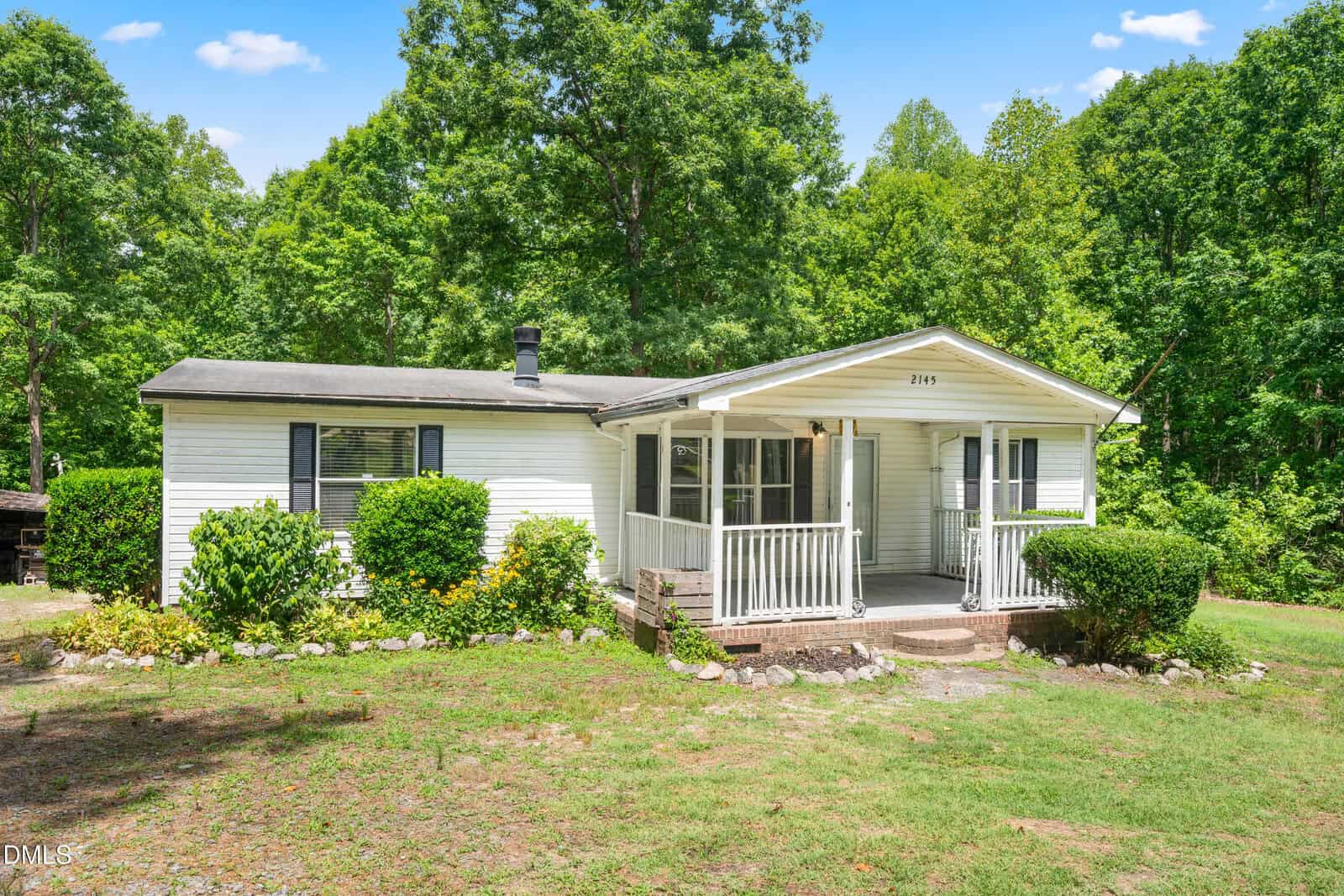 A small white house with a covered front porch, black shutters, and a gently sloped roof sits surrounded by green bushes, grass, and trees under a blue sky with scattered clouds.