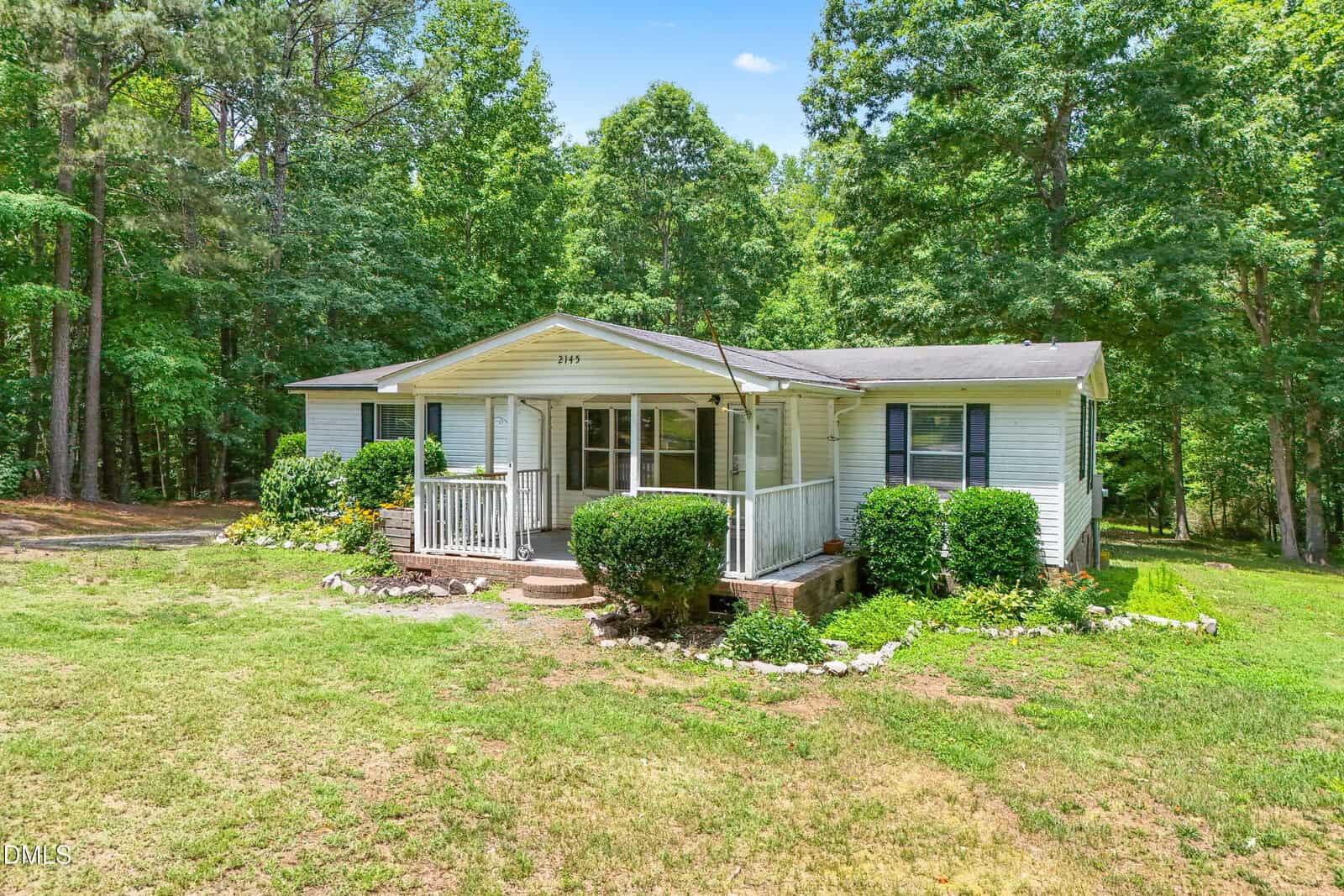 A single-story white house at 2145 Antler Way Lane in Creedmoor features a small front porch with steps, surrounded by green bushes and trees on a grassy lawn under a blue sky.