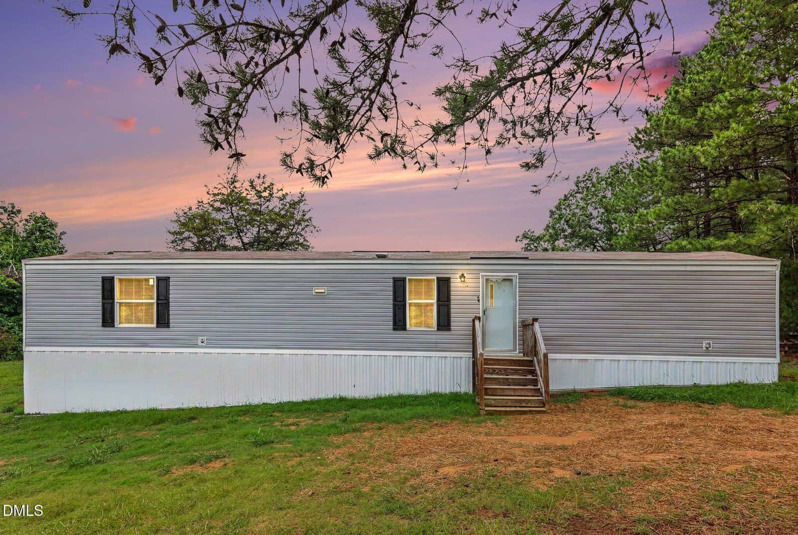 A single wide gray mobile home with white trim and black shutters sits on a grassy lawn at 20 Christian Life Lane in Louisburg, surrounded by trees, under a colorful sunset sky. Wooden steps lead to the front door.