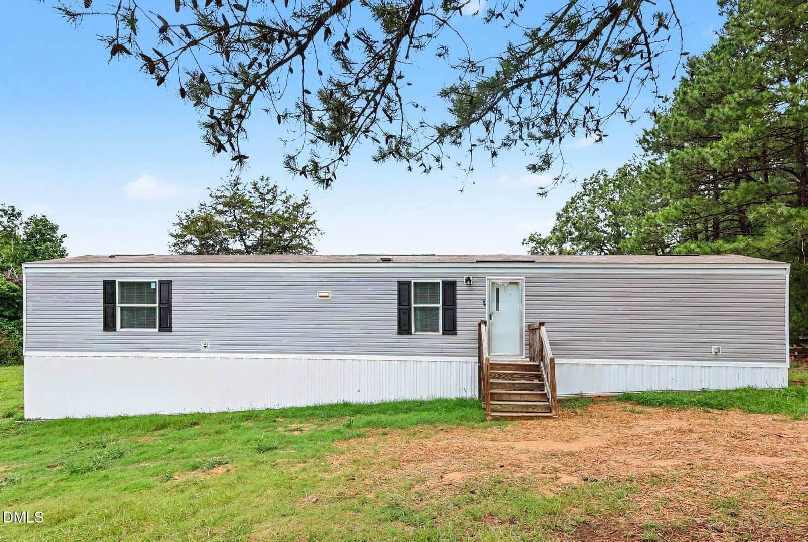 A light gray mobile home with white trim and two windows sits on a grassy lot with trees in the background. Wooden steps lead to the front door under a partly cloudy sky.