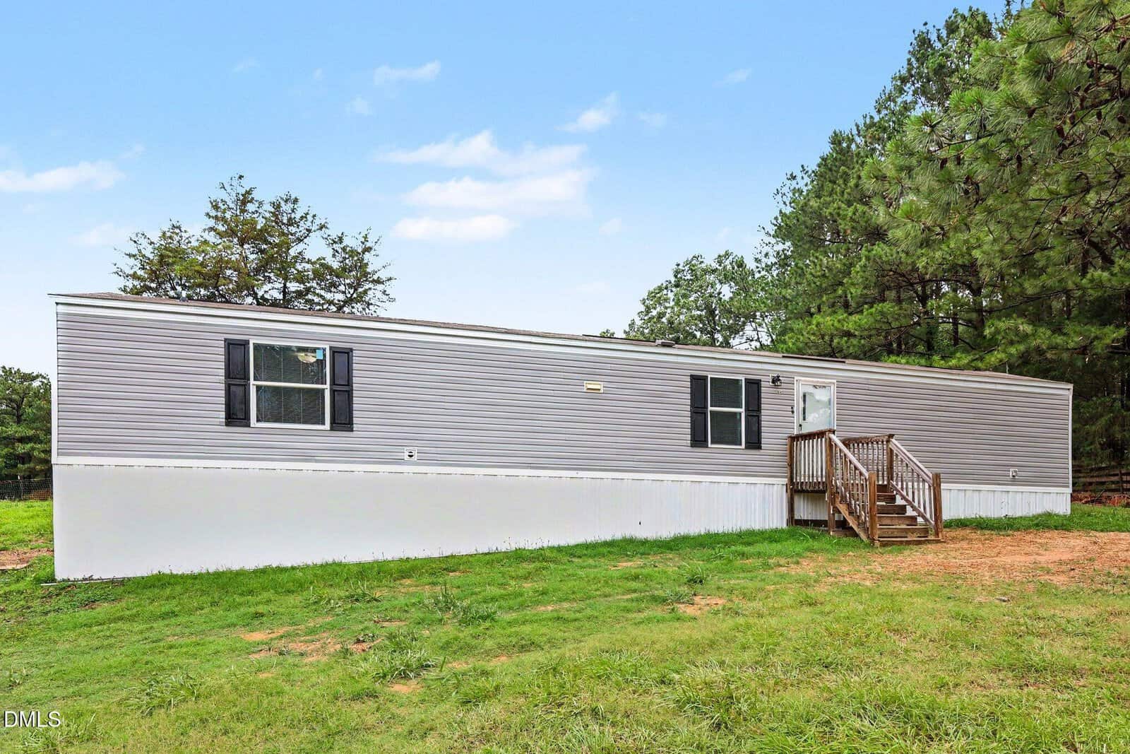 A beige mobile home with black shutters sits on a grassy lot, bordered by trees. Wooden steps lead up to the front door under a clear blue sky.