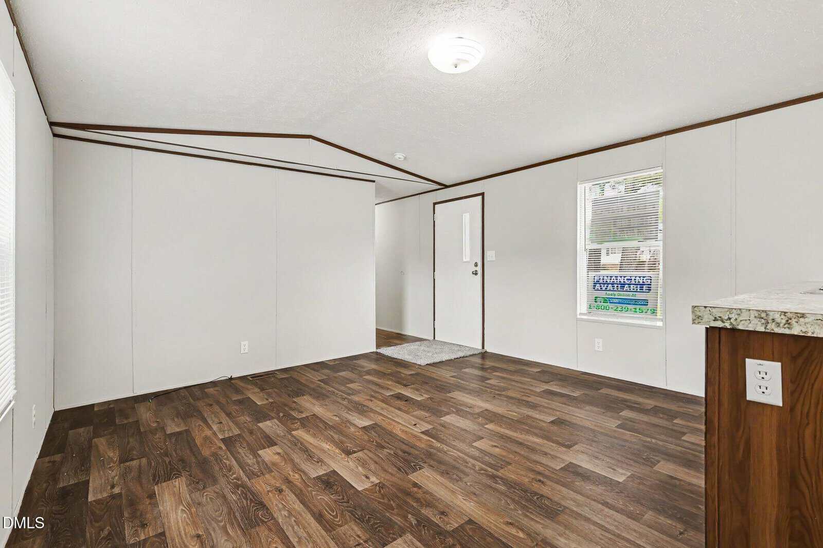 A spacious, empty room with white walls, dark wood flooring, a small light fixture on the ceiling, and a white front door next to a window with a "Financing Available" sign outside.