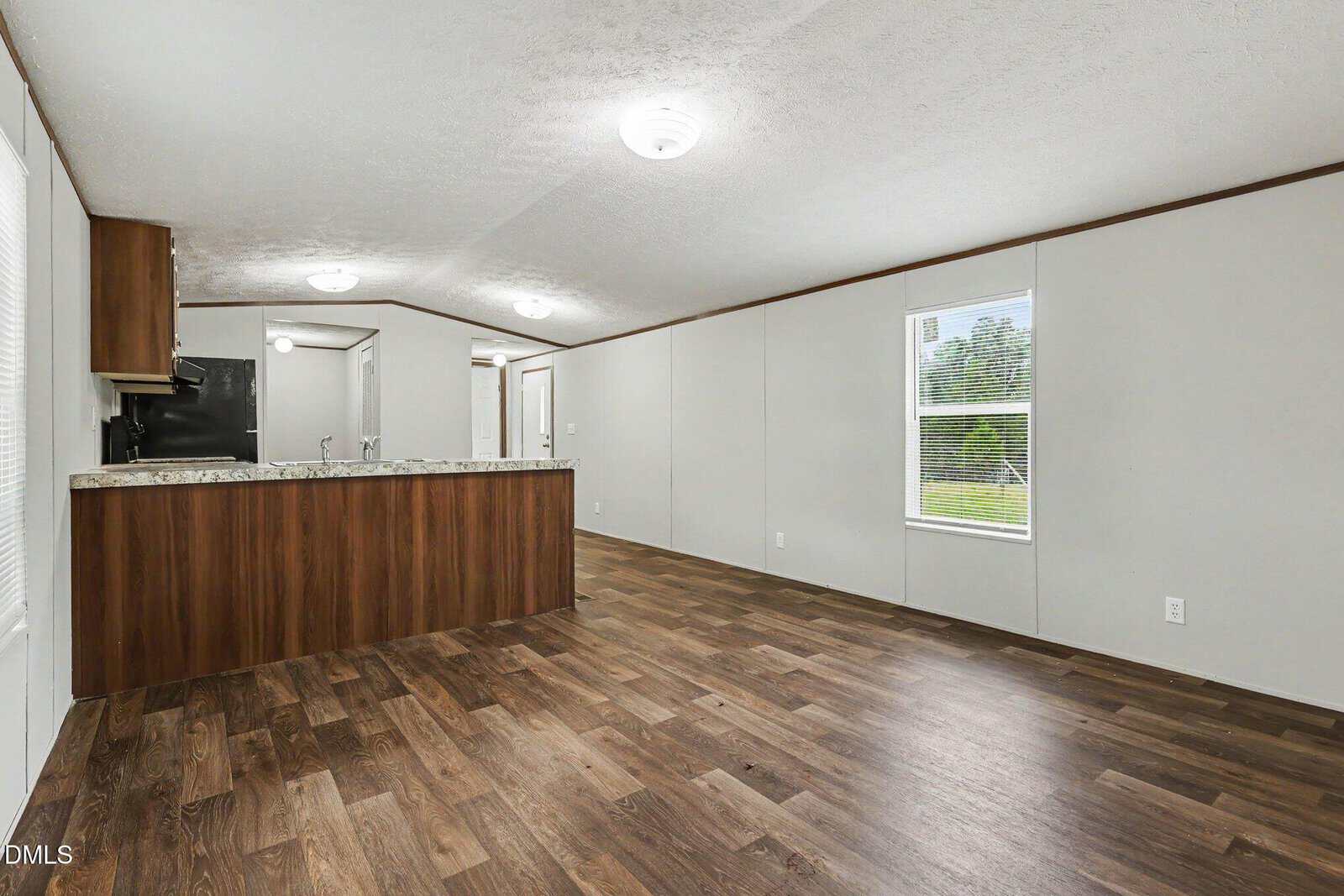 An empty room with wood-look flooring, off-white walls, a large window with blinds, ceiling lights, and an open kitchen area with wood cabinets and a marble-style countertop.