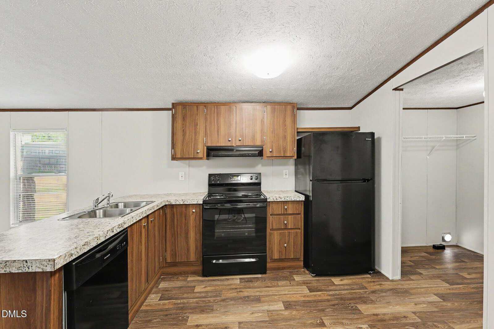 Modern kitchen with wood cabinets, black appliances, light countertops, double sink, and wood-style flooring. A pantry with an open door is visible on the right. Natural light enters from a window on the left.