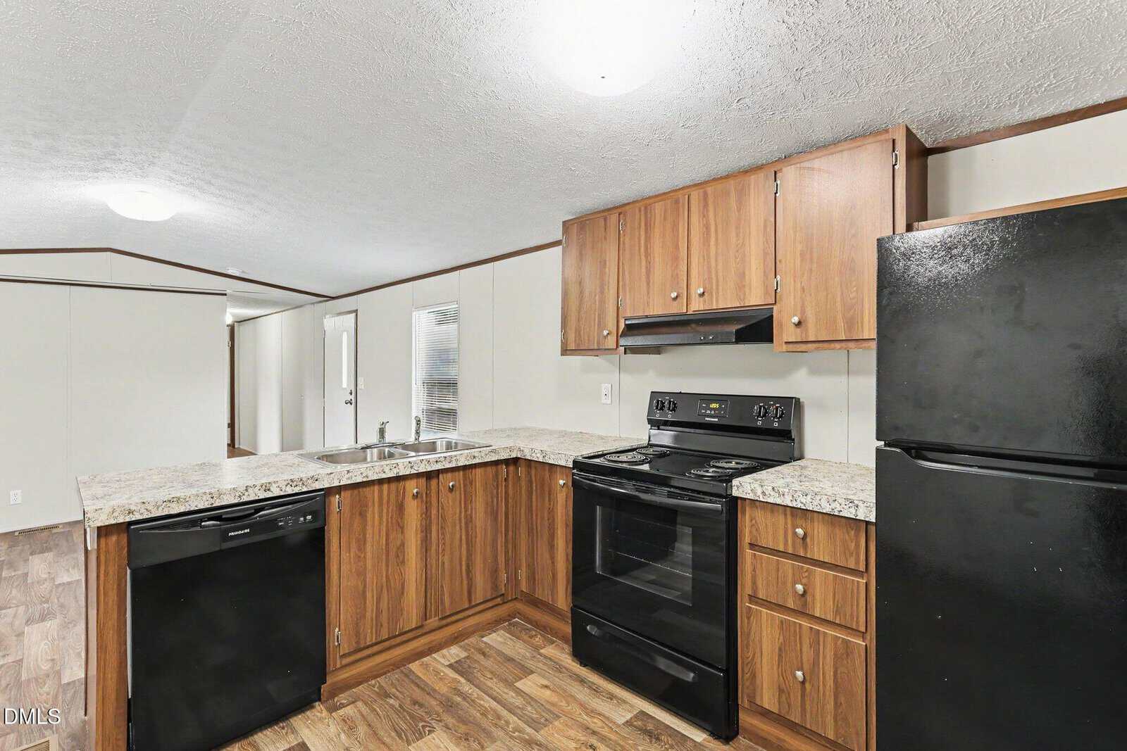 Modern kitchen with wood cabinets, black appliances including a stove, dishwasher, and refrigerator, granite countertops, and a double sink. The space opens to a hallway and door with ample natural light.