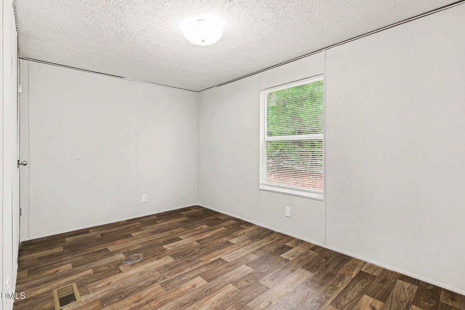 Empty room with light gray walls, wood-look vinyl flooring, a white ceiling light, a window with blinds, and a closed white door. Ceiling has a subtle textured finish.