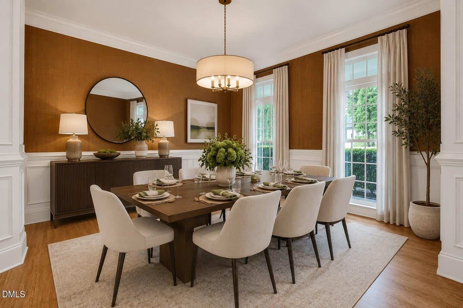 Elegant dining room with a wooden table set for eight, cream upholstered chairs, a beige rug, large windows with white curtains, a round mirror, lamps, and floral decor on a sideboard against a brown accent wall.