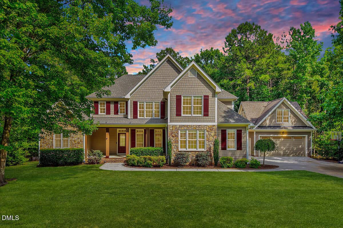 A two-story suburban house at 1006 Chatham Court in Youngsville features a stone and siding exterior, red shutters, covered porch, manicured lawn, double garage, and is surrounded by lush trees under a colorful sunset sky.
