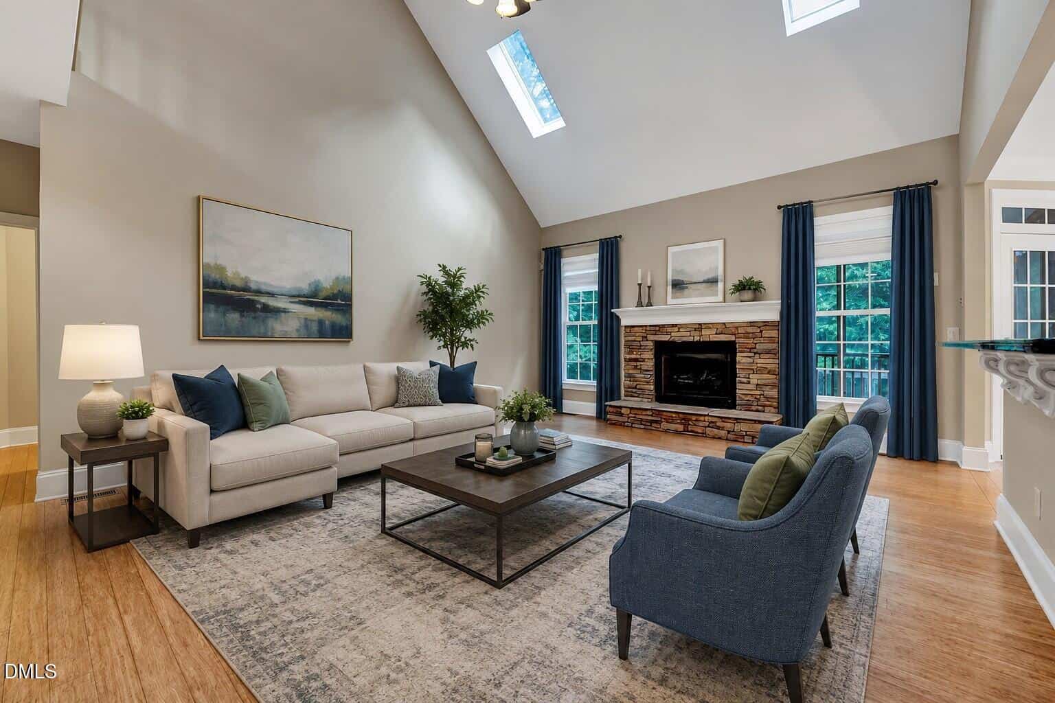 A modern living room with a beige sofa, two blue armchairs, a stone fireplace, a large painting, blue curtains, skylights, and a coffee table on a patterned rug. Light streams in from windows and skylights.