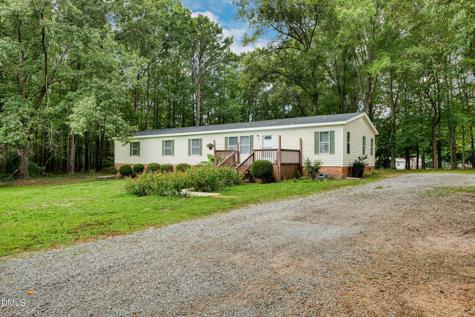 A single-story light yellow house with a front porch and steps, surrounded by trees and greenery, with a gravel driveway in the foreground.