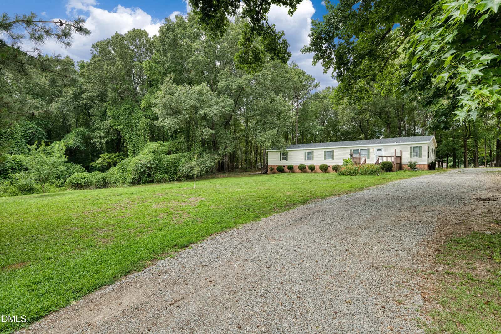 A single-story white house with green shutters sits in a large grassy yard, surrounded by tall trees. A gravel driveway curves toward the house, and the sky above is partly cloudy.