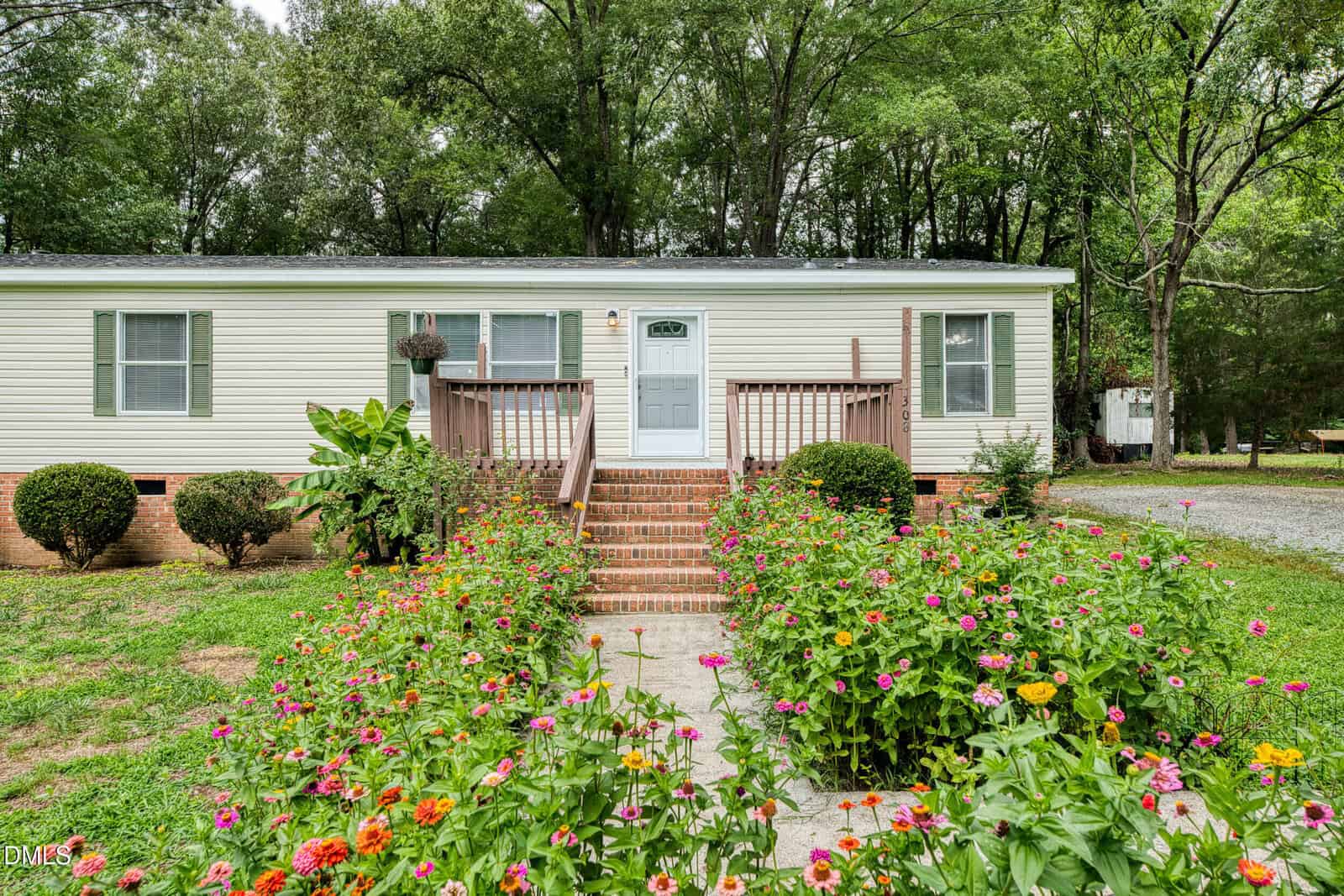 A single-story white house with a front porch and steps, surrounded by blooming flower beds and lush greenery, set against a backdrop of tall trees.