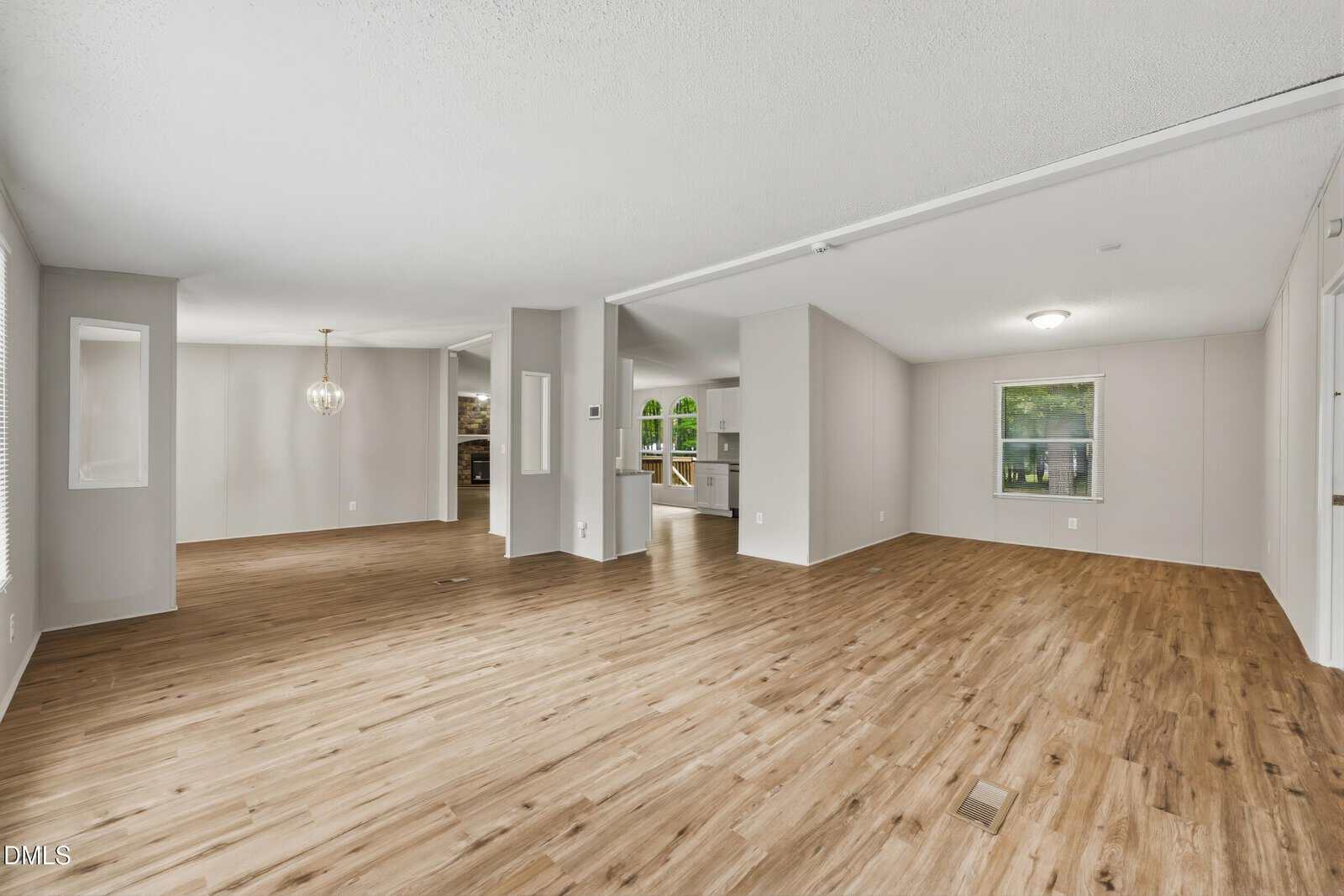 Spacious open-concept living and dining area with light wood flooring, neutral walls, large windows, and a modern light fixture. The kitchen is visible in the background.