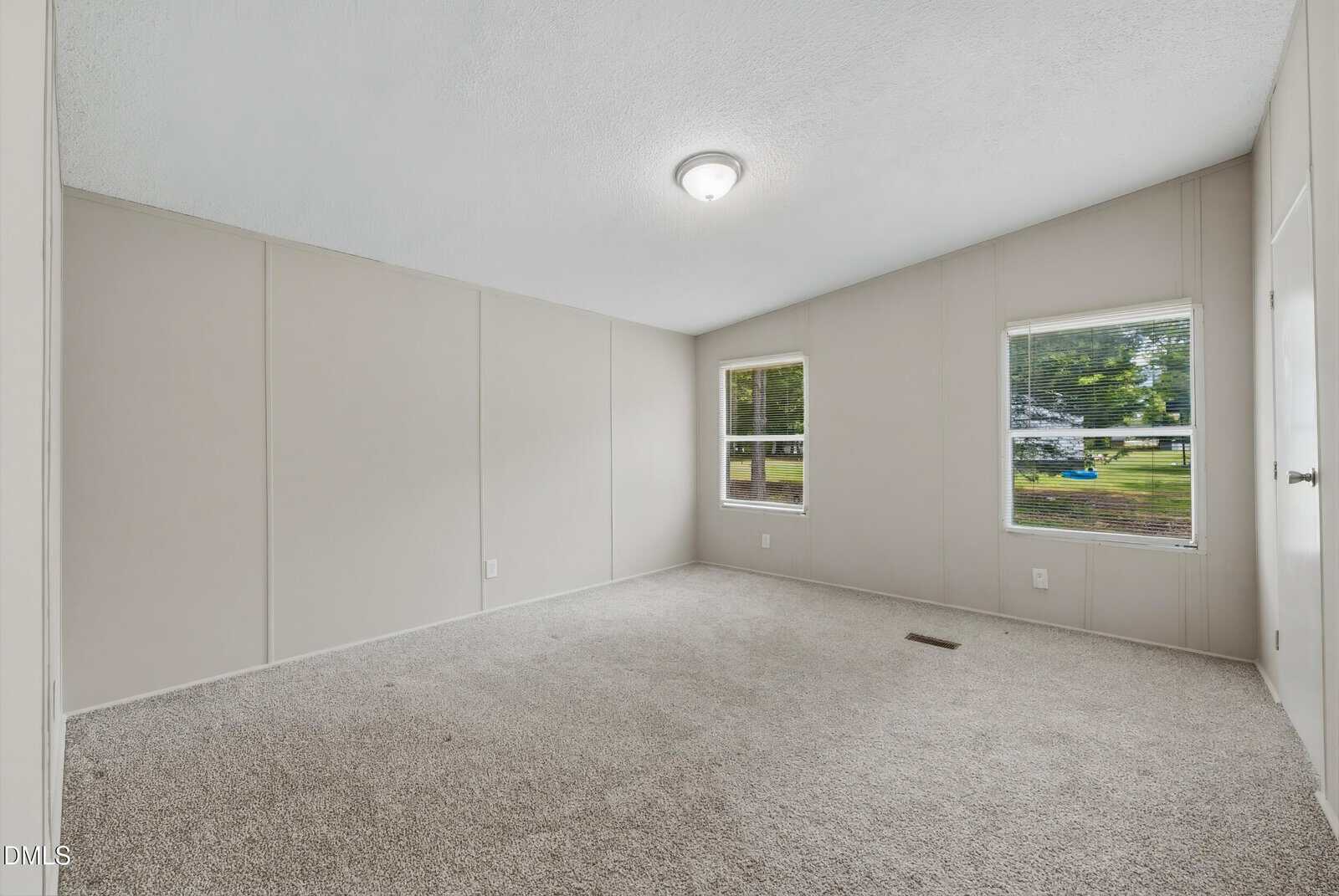 Empty room with beige walls, white ceiling, light fixture, two windows letting in natural light, and light gray carpeted flooring. Some grass and trees are visible outside the windows.