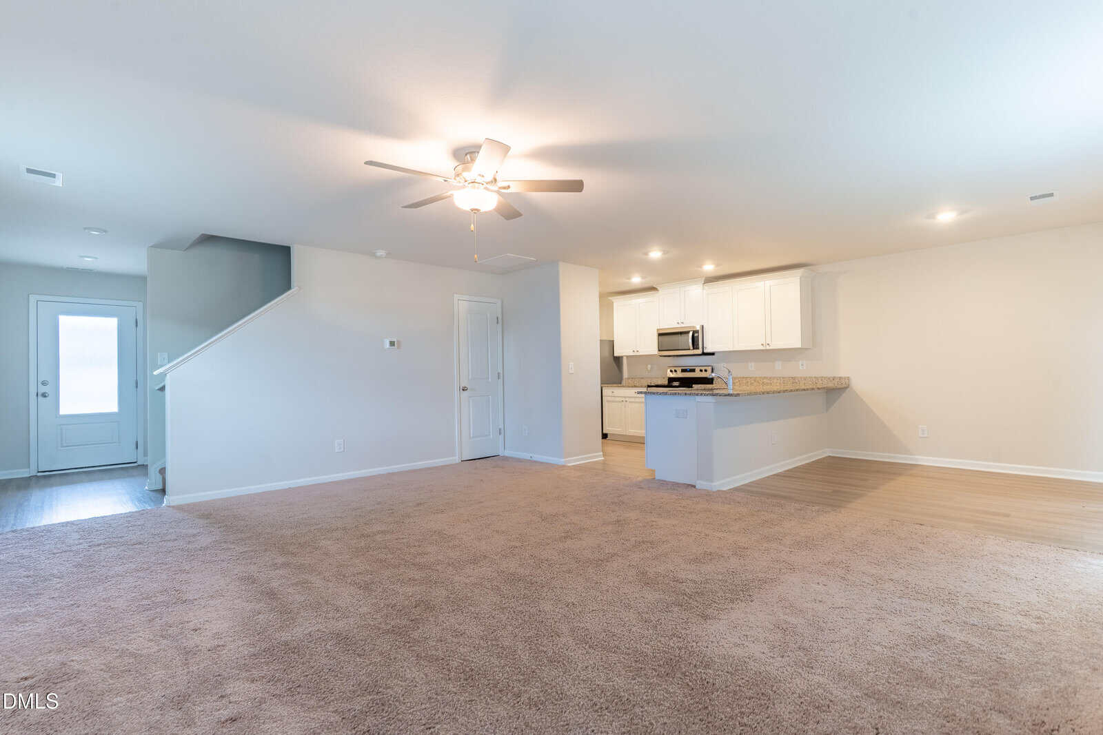 Open living area with beige carpet, ceiling fan, and adjacent kitchen featuring white cabinets and stainless steel appliances; stairs and a back door are visible to the left.