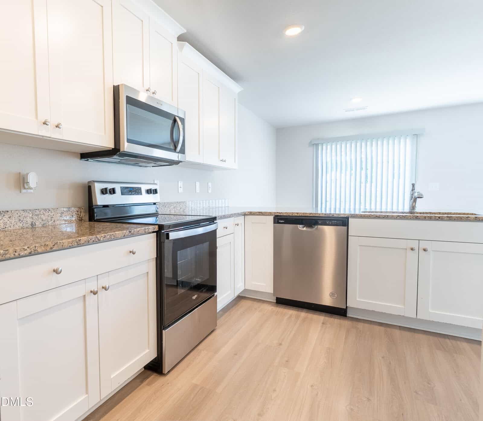 Modern kitchen with white cabinets, stainless steel appliances including a stove, microwave, and dishwasher, granite countertops, light wood flooring, and a window with vertical blinds above the sink.
