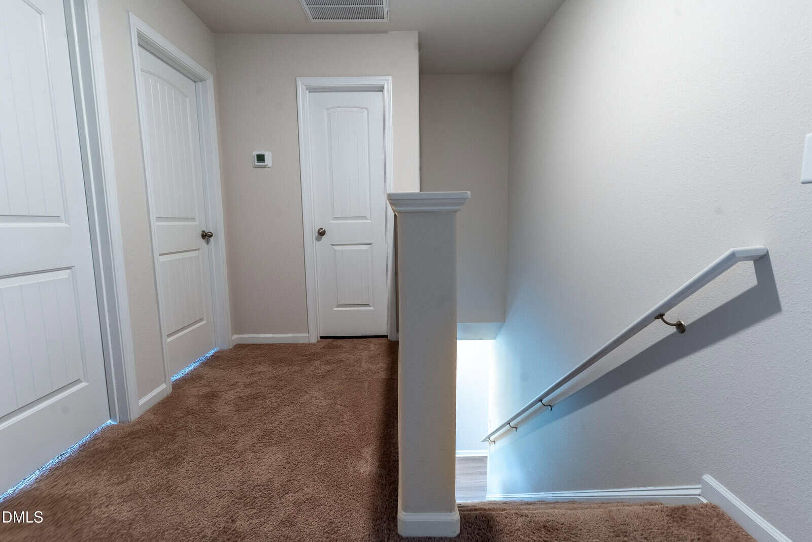 A carpeted hallway with three closed white doors and a stairway with a handrail leading down. The walls are light-colored, and there is a thermostat on one wall.