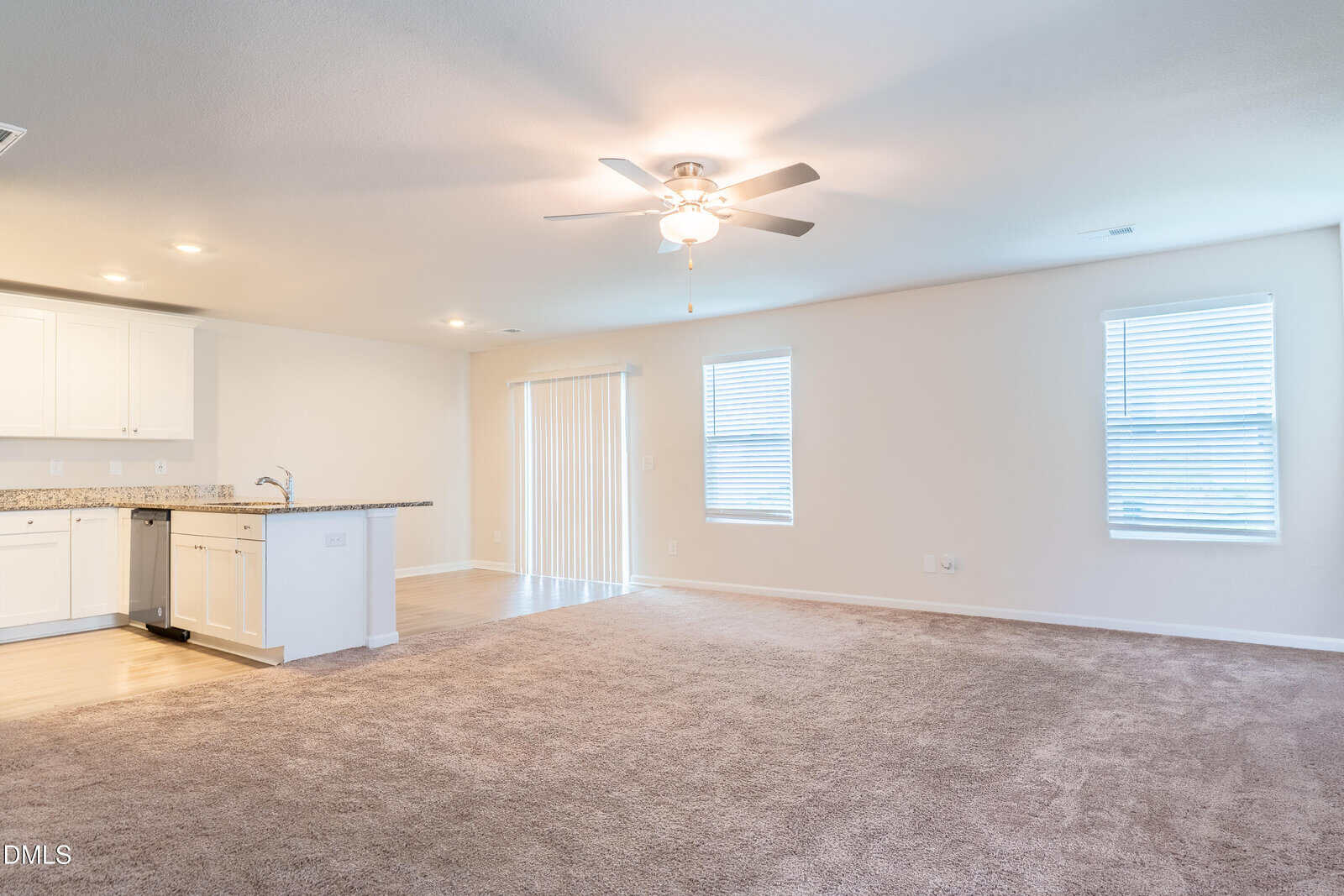 Spacious, empty living room with beige carpet, white walls, and two windows. Ceiling fan with light is on. Open kitchen with white cabinets and granite countertops is visible to the left. Sliding glass doors lead outside.