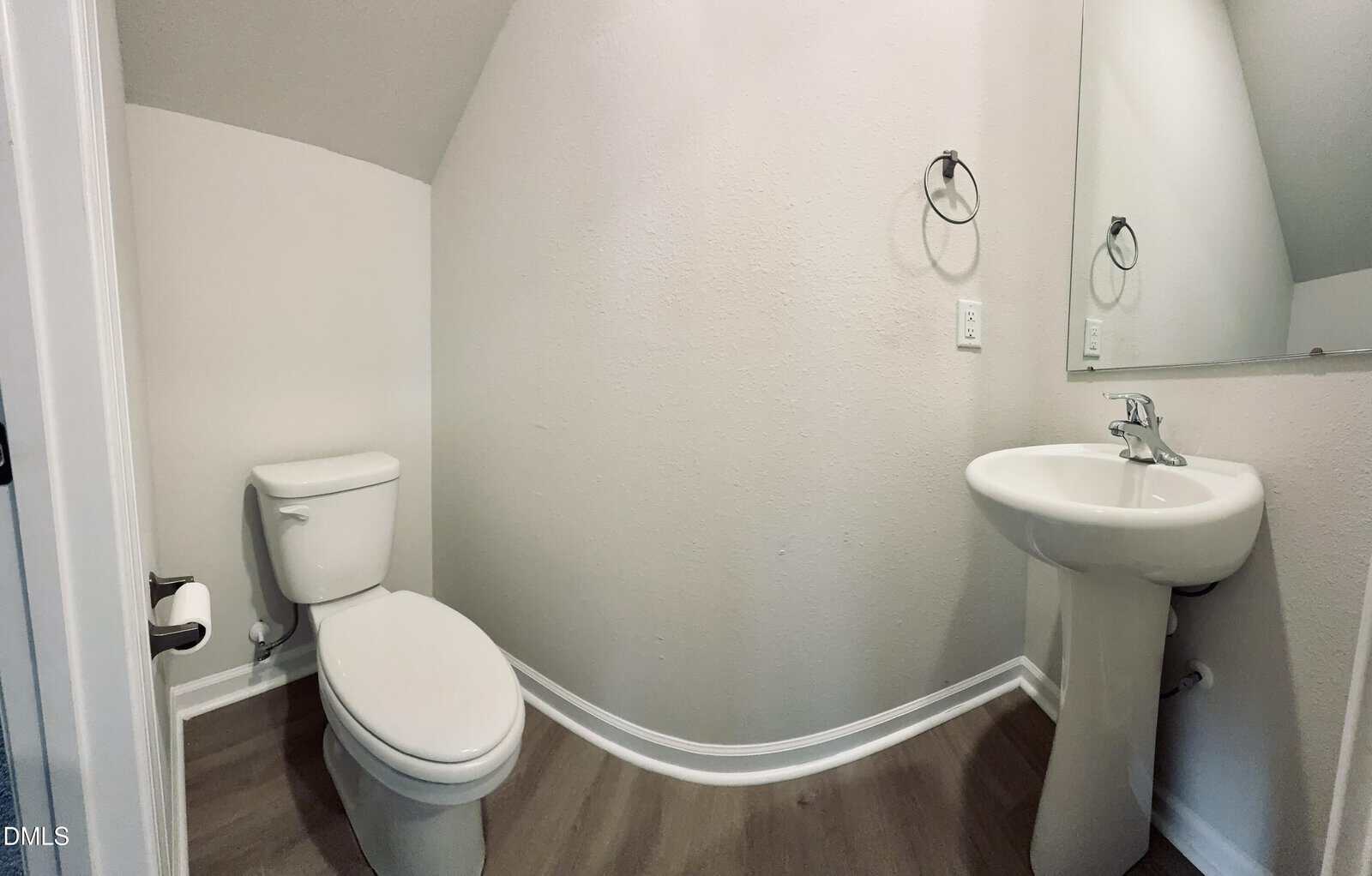 A small bathroom with light-colored walls, a white toilet, a pedestal sink, a wall mirror, and towel rings. The floor is wood-style, and the room has an angled ceiling.