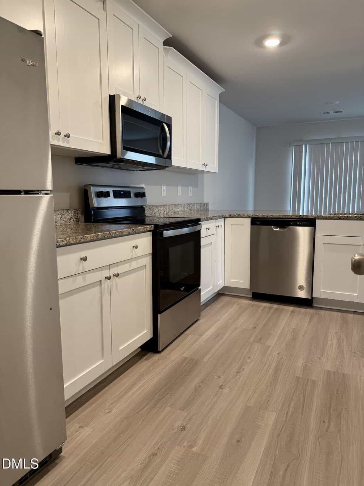 Modern kitchen with stainless steel appliances, including a refrigerator, oven, microwave, and dishwasher. White cabinets, granite countertops, and light wood flooring. Vertical blinds cover a large window in the background.