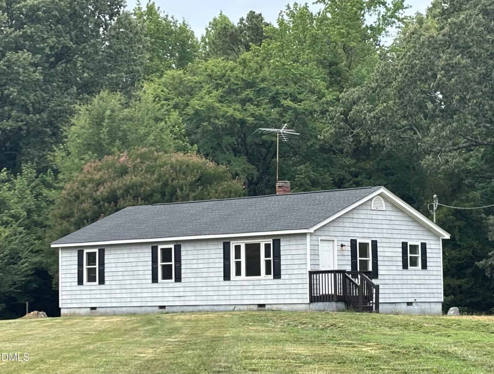 A small, single-story gray house with white trim sits on a grassy lawn at 695 Layton Store Road in Castalia, surrounded by dense green trees. The house has several windows and a small front porch with steps leading up to the door.