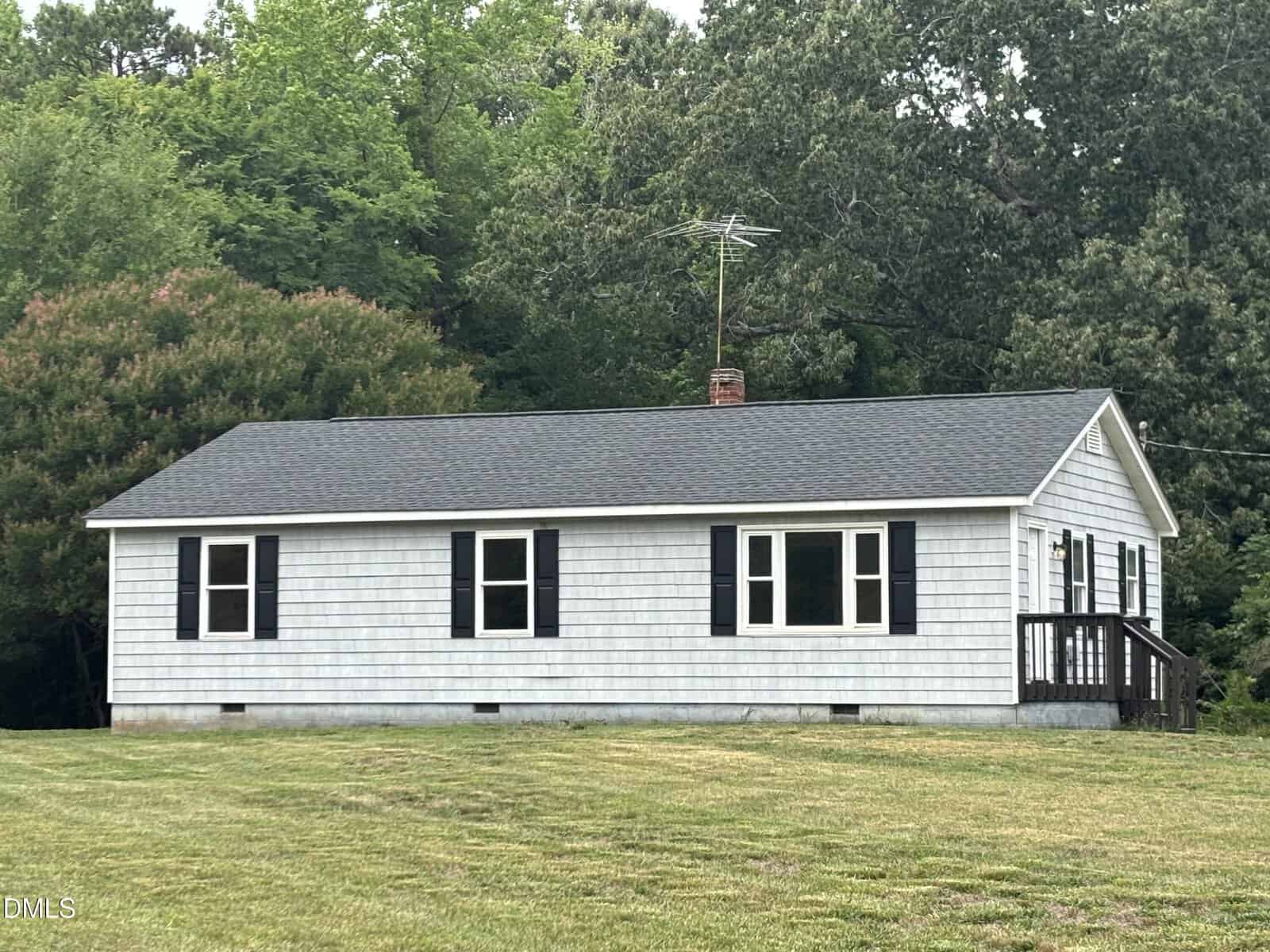 A small, single-story gray house with black shutters and a dark roof sits on a grassy lawn, surrounded by green trees. The house has a front porch with steps and an antenna on the roof.