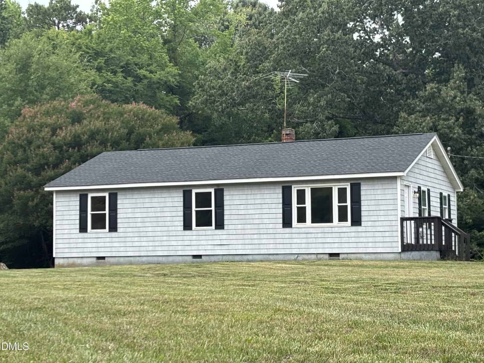 A small, single-story house with gray siding, black shutters, and a dark roof sits on a large grassy lawn. Trees and greenery surround the house, and there is a small porch on the right side.