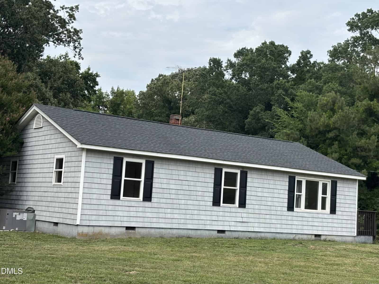 A single-story gray house with black shutters, shingle siding, and a dark roof sits on a grassy lawn, surrounded by tall, leafy trees in the background.