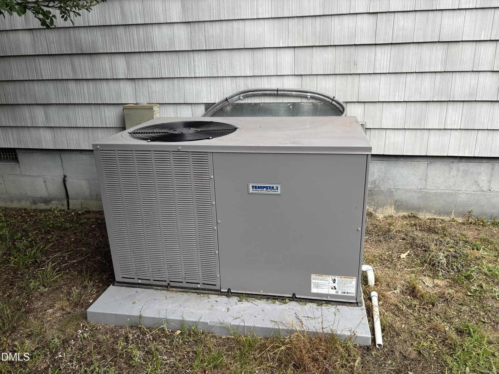 A Tempstar HVAC unit sits on a concrete pad next to a house with gray shingle siding, surrounded by grass and dirt.