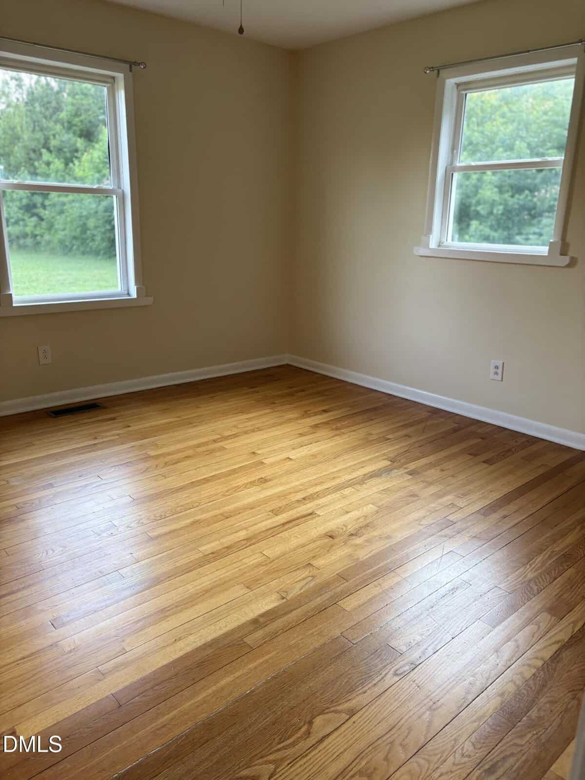 Empty room with light-colored walls, two windows with views of green trees, and a polished hardwood floor. Daylight brightens the space, and there are electrical outlets on two walls.