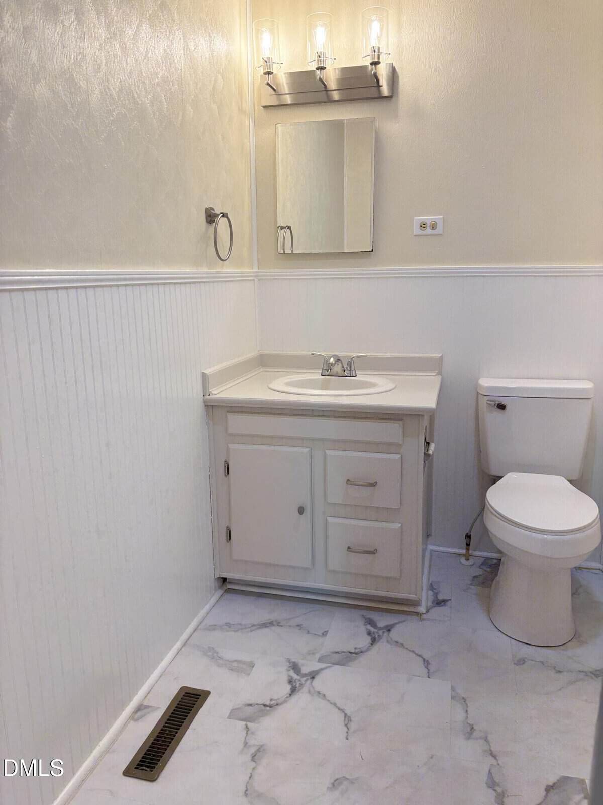 A small bathroom with a white vanity, sink, mirror, wall-mounted lights, toilet, and marble-patterned floor. The walls are light with beadboard wainscoting.