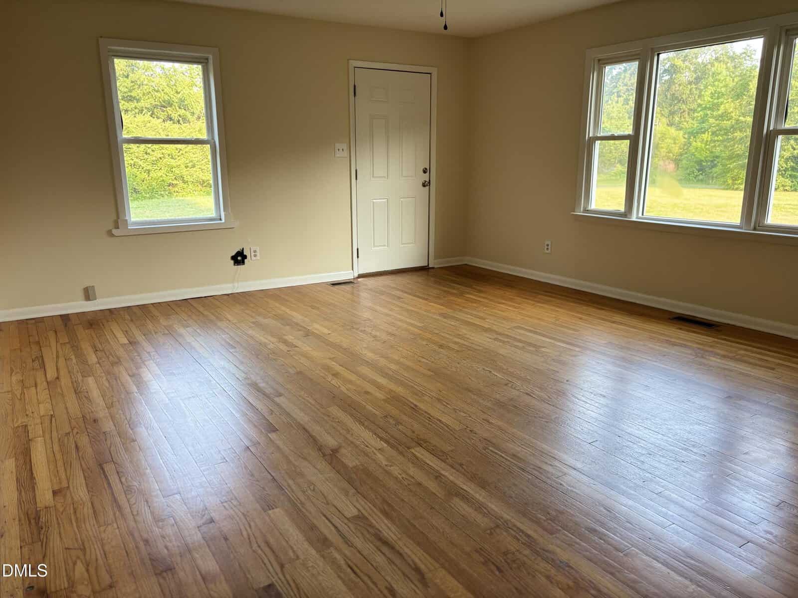 Empty room with hardwood floor, beige walls, two large windows showing green trees outside, and a white door. Sunlight streams in, creating a bright and clean atmosphere.