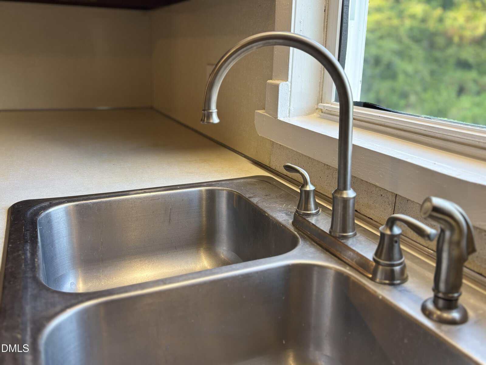 Close-up of a stainless steel double kitchen sink with a silver faucet and handles, set against a beige countertop and backsplash, next to a window with greenery visible outside.