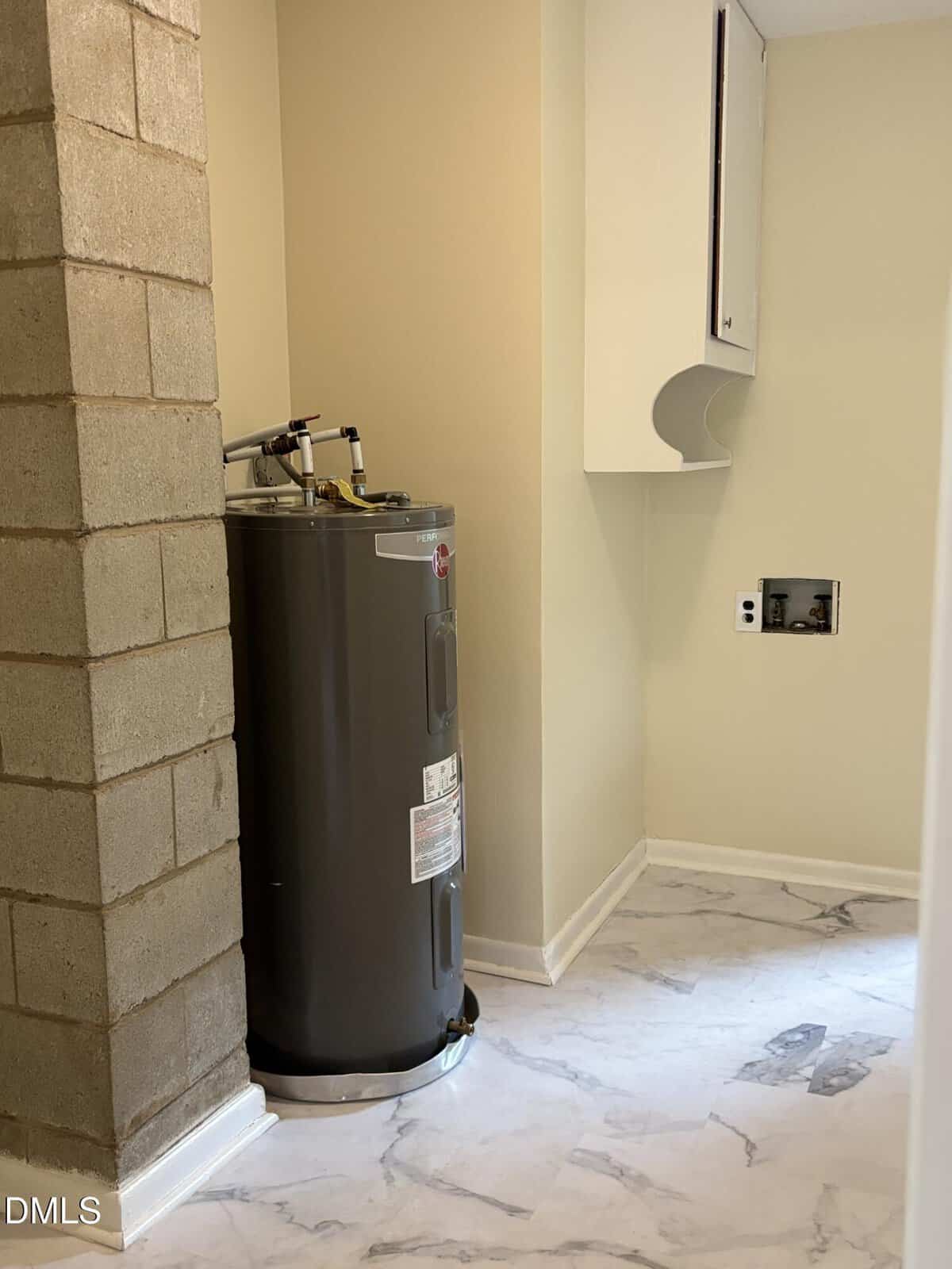 A gray water heater stands in a corner next to a cinder block wall in a laundry room with white cabinets, beige walls, and white marble-patterned floor tiles.