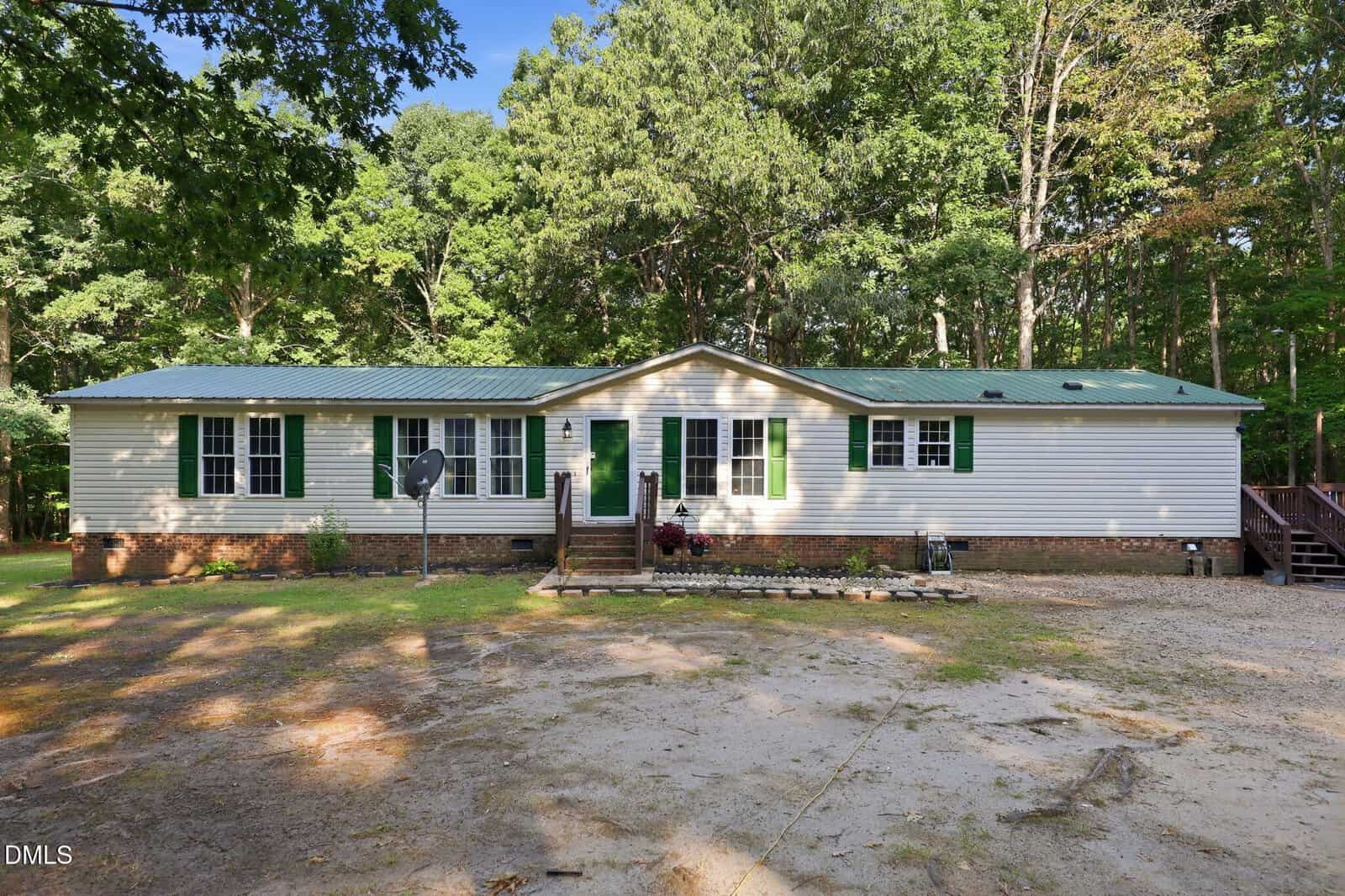 A single-story white house with green shutters and a green metal roof, surrounded by tall trees. There are steps leading up to a central front door and a satellite dish on the left side of the yard.