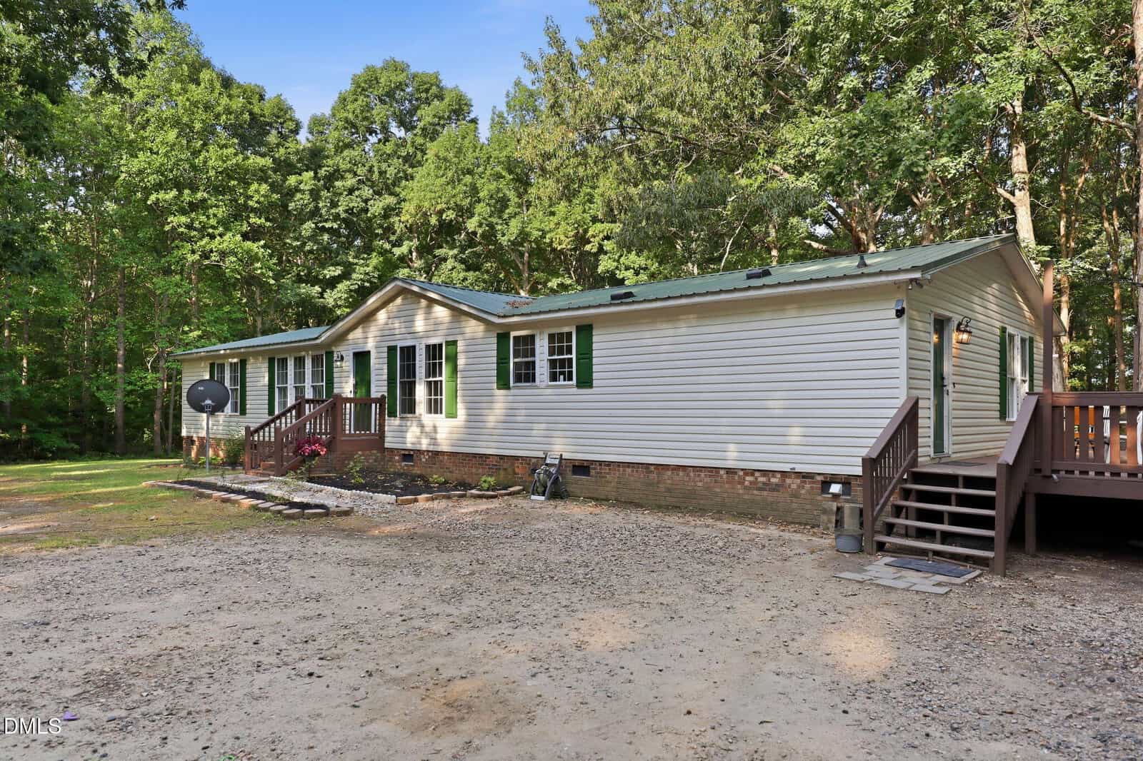 Single-story beige manufactured home with green shutters, front steps, small garden, and a gravel driveway, surrounded by trees and greenery under a clear blue sky.