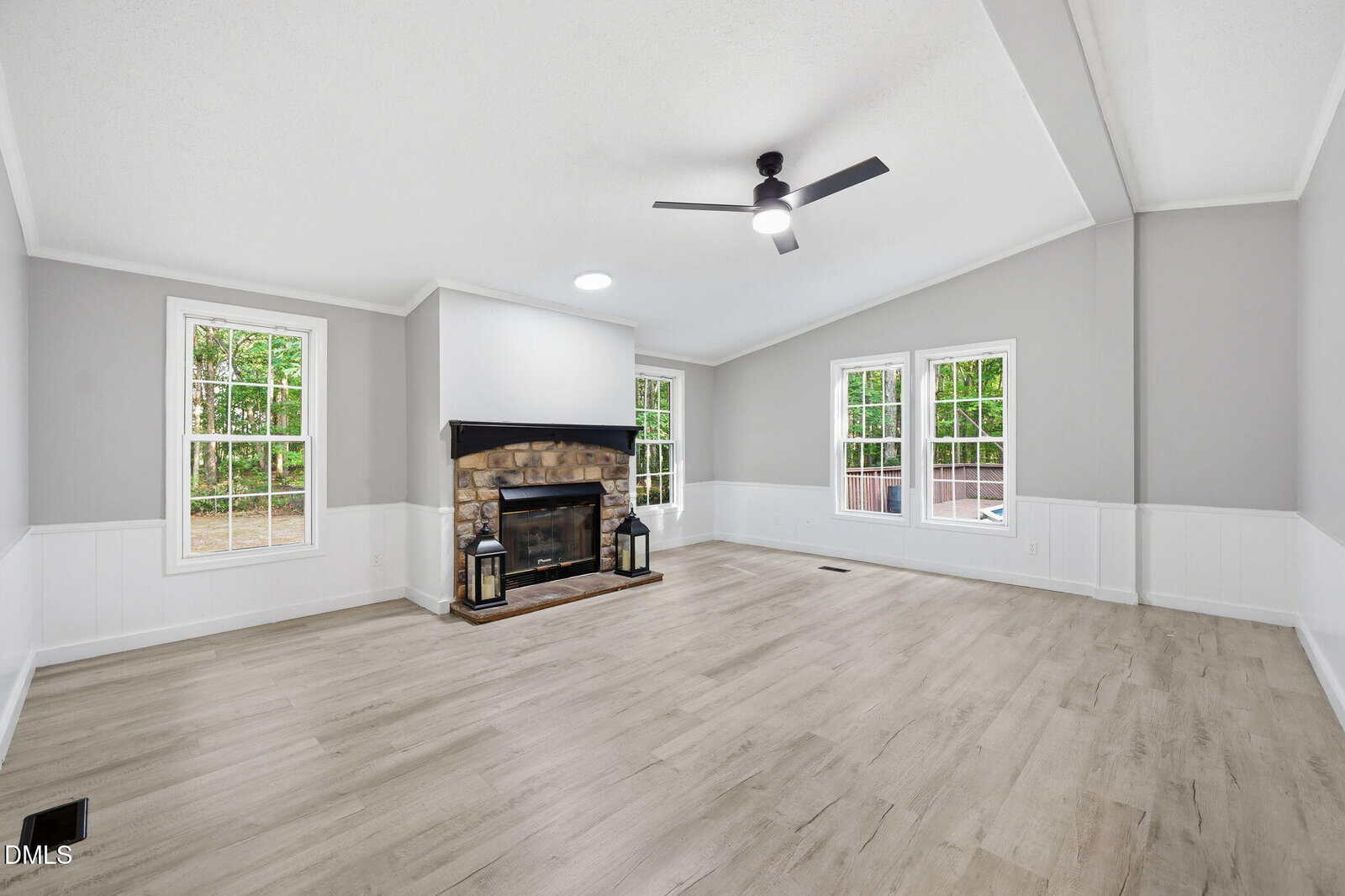 Spacious living room with light wood floors, gray and white walls, large windows, and a stone fireplace with two lanterns. Ceiling fan with light above.